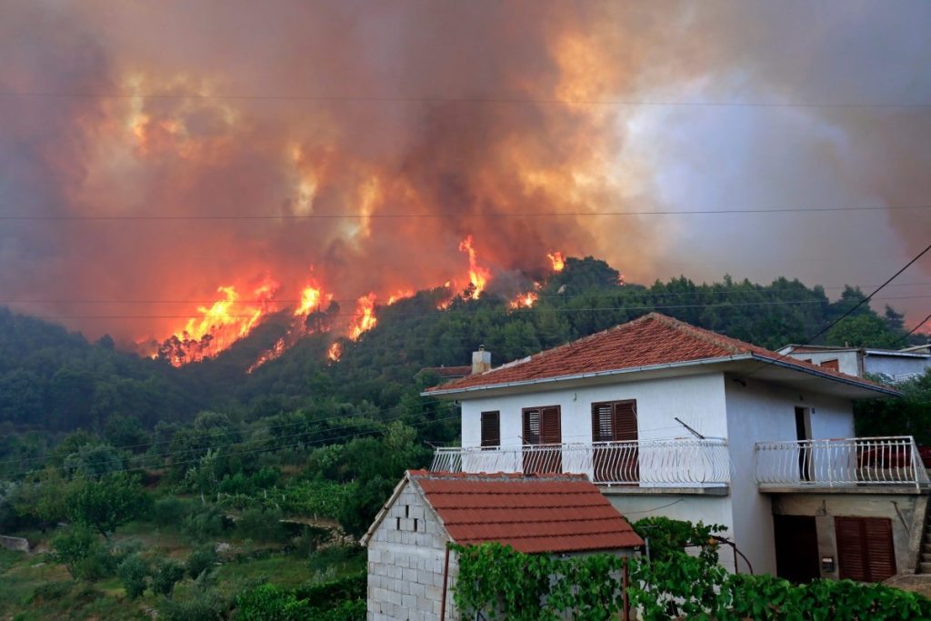 A fire burns near a house in Greece, raising concerns for fire safety inspection and the need for home hardening measures.